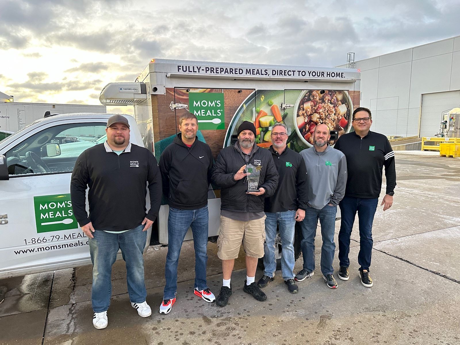 Six Mom’s Meals team members standing in front of a branded delivery truck, with Ken, the first recipient of the Mom’s Hero Award, holding his award.